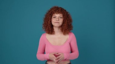 Positive curly haired redhead woman wearing pink shirt agrees with something in blue studio