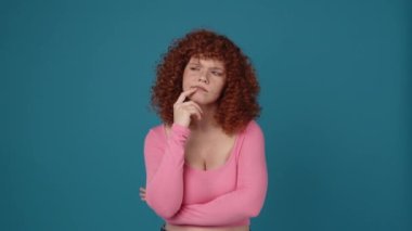 Meditasyon yapan kıvırcık saçlı, pembe tişört giyen kızıl saçlı kadın mavi stüdyodaki bir şeye katılmıyor.