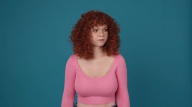 Sad curly haired redhead woman wearing pink shirt screaming at the camera in blue studio