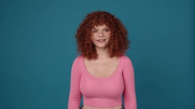 Surprised curly haired redhead woman wearing pink shirt looking at the camera in blue studio