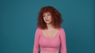 Upset curly haired redhead woman wearing pink shirt looking around in blue studio