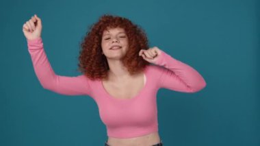 Funny curly haired redhead woman wearing pink shirt dancing in blue studio