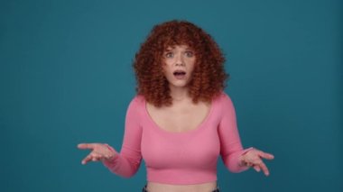 Shocked curly haired redhead woman wearing pink shirt looking at the camera in blue studio