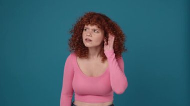 Pretty curly haired redhead woman wearing pink shirt shows she doesn't hear anything in blue studio