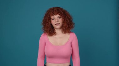 Displeased curly haired redhead woman wearing pink shirt looking at the camera in blue studio