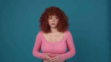 Not understanding curly haired redhead woman wearing pink shirt spreading hands in blue studio