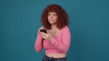 Pensive curly haired redhead woman wearing pink shirt texting on phone in blue studio