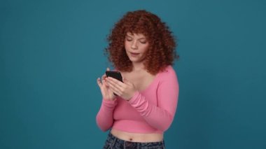 Smiling curly haired redhead woman wearing pink shirt typing on phone in blue studio
