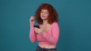 Meditative curly haired redhead woman wearing pink shirt texting on mobile in blue studio