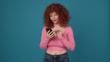 Smiling curly haired redhead woman wearing pink shirt talking by video call on phone in blue studio