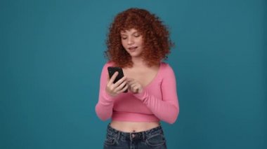 Cheerful curly haired redhead woman wearing pink shirt talking by video call on phone in blue studio
