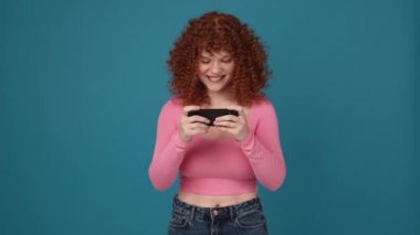 Cheerful curly haired redhead woman wearing pink shirt playing digital game on phone in blue studio