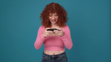 Happy curly haired ginger woman wearing pink shirt playing digital game on phone in blue studio