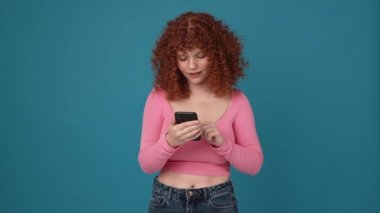Positive curly haired ginger woman wearing pink shirt making selfie on mobile in blue studio