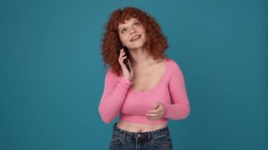 Mavi stüdyoda pembe tişörtlü mutlu kıvırcık saçlı kadın telefonla konuşuyor.