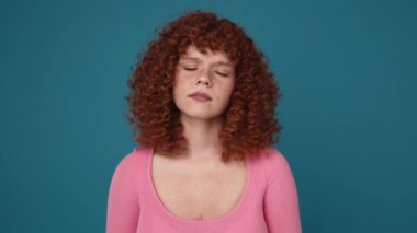 Bored curly haired ginger woman wearing pink shirt yawning in blue studio