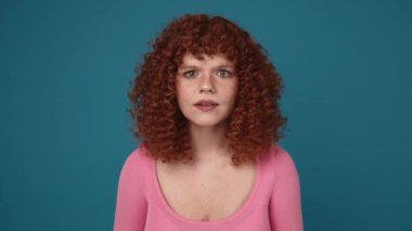 Surprised curly haired ginger woman wearing pink shirt peering at the camera and spreading hands in blue studio