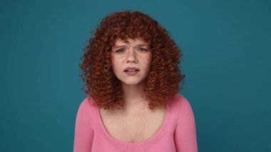 Displeased curly haired ginger woman wearing pink shirt peering at the camera and smiling in blue studio