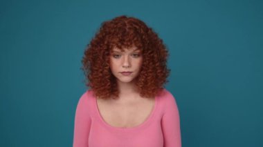 Pretty curly haired ginger woman wearing pink shirt looking at the camera in blue studio
