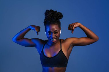 Black young sportswoman in earphones showing her biceps isolated over blue background