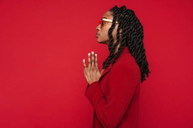 Kırmızı arka planda izole edilmiş bir şekilde el kol hareketi yaparken ceketini giyen siyah genç bir kadın yan tarafa bakıyor.
