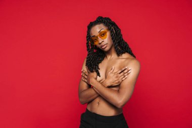 Black shirtless woman in sunglasses posing and covering her bust isolated over red background