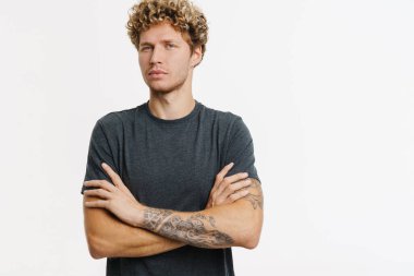 Sarışın genç adam beyaz duvarın üzerinde kollarını kavuşturmuş poz verirken kameraya bakıyor.