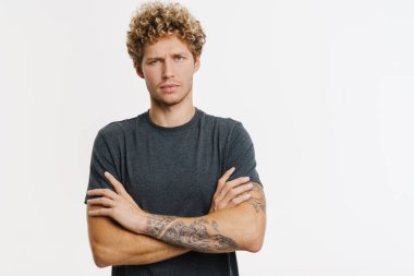 Sarışın genç adam beyaz duvarın üzerinde kollarını kavuşturmuş poz verirken kaşlarını çatıyor.