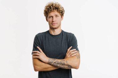 Sarışın genç adam beyaz duvarın üzerinde kollarını kavuşturmuş poz verirken kaşlarını çatıyor.