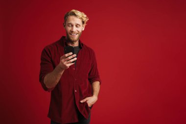 Kırmızı duvardan izole edilmiş, gülümseyen ve cep telefonu kullanan beyaz sakallı bir adam.