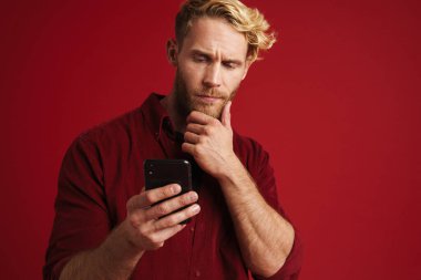 Kırmızı duvardan izole edilmiş cep telefonu kullanan beyaz şaşkın bir adam.