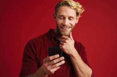 Kırmızı duvardan izole edilmiş, gülümseyen ve cep telefonu kullanan beyaz sakallı bir adam.