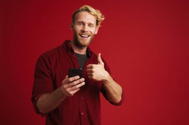 Sakallı sarışın adam baş parmağını gösteriyor ve kırmızı arka planda izole edilmiş cep telefonu kullanıyor.