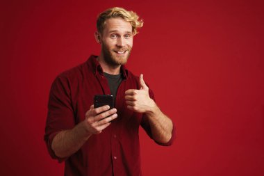 Sakallı sarışın adam baş parmağını gösteriyor ve kırmızı arka planda izole edilmiş cep telefonu kullanıyor.