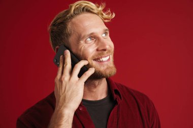 Sakallı sarışın adam kırmızı arka planda izole edilmiş, gülümseyen ve cep telefonuyla konuşan.