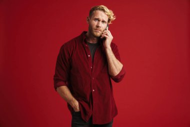 Beyaz sakallı adam kırmızı duvarda izole bir şekilde cep telefonuyla konuşurken kaşlarını çatıyor.