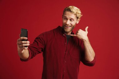 Beyaz sakallı adam kırmızı duvardan izole edilmiş cep telefonuyla selfie çekerken çağrı hareketi yapıyor.