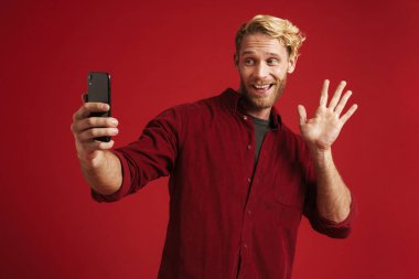Beyaz sakallı adam kırmızı duvarda izole edilmiş cep telefonuyla selfie çekerken el sallıyor.
