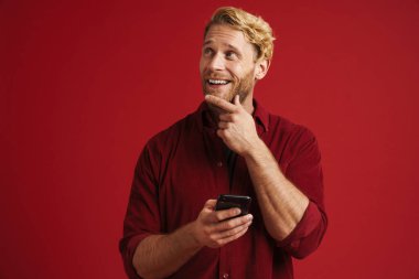 Kırmızı duvardan izole edilmiş, gülümseyen ve cep telefonu kullanan beyaz sakallı bir adam.