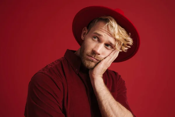 White bearded man wearing hat propping up his head and looking aside isolated over red wall