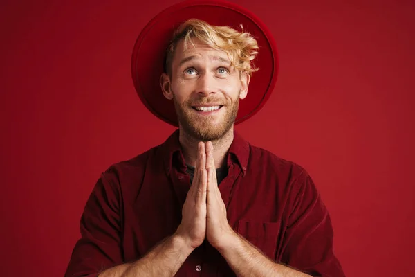 White bearded man wearing hat smiling while making prayer gesture isolated over red wall