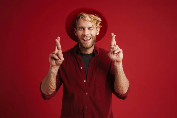 White bearded man wearing hat holding fingers crossed for good luck isolated over red wall