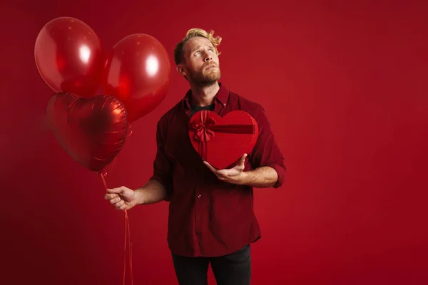 White bearded man posing with balloons and heart gift box isolated over red wall
