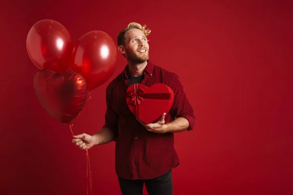 Bearded blonde man smiling while posing with balloons and gift box isolated over red background