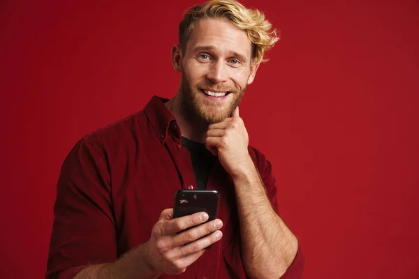 Bearded blonde man smiling and using mobile phone isolated over red background