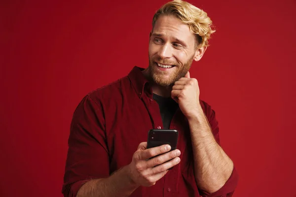 Bearded blonde man smiling and using mobile phone isolated over red background