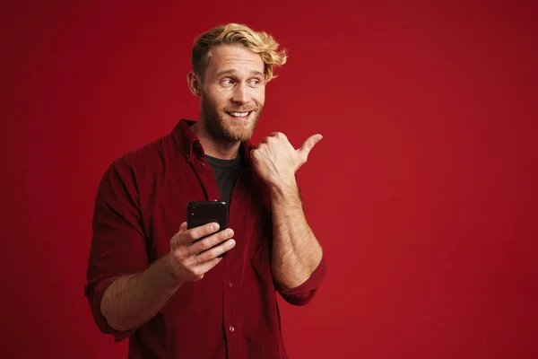 Bearded blonde man pointing finger aside and using mobile phone isolated over red background