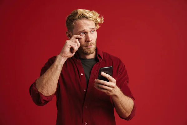 White bearded man crying while using mobile phone isolated over red wall