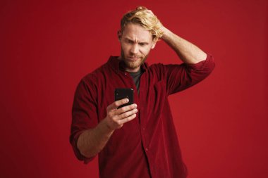 Kırmızı duvardan izole edilmiş cep telefonu kullanan beyaz şaşkın bir adam.