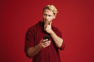 Kırmızı duvardan izole edilmiş cep telefonu kullanan beyaz şaşkın bir adam.
